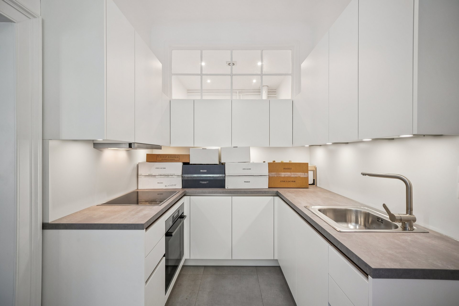 Modern U-shaped kitchen with white cabinets, grey countertops, stainless steel sink, and cardboard boxes on the counter. [AI-generated text]