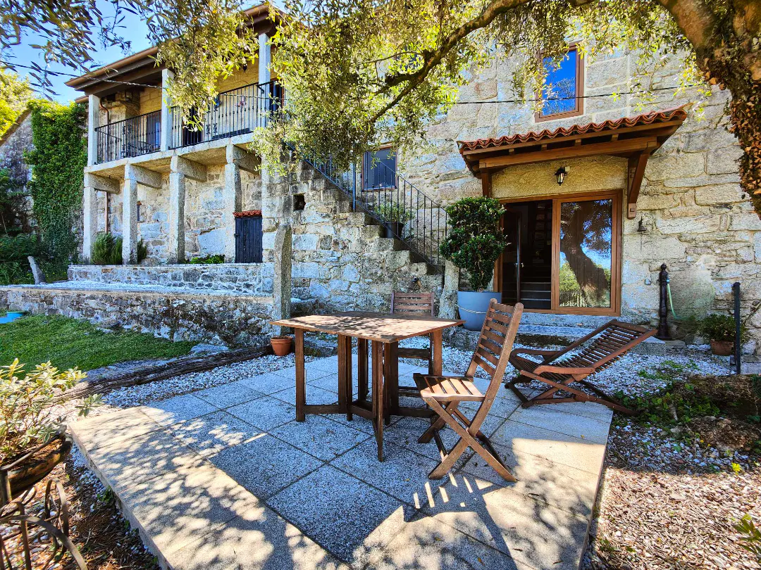 Rustic stone house with a patio, wooden table and chairs, and an outdoor staircase under a tree on a sunny day.