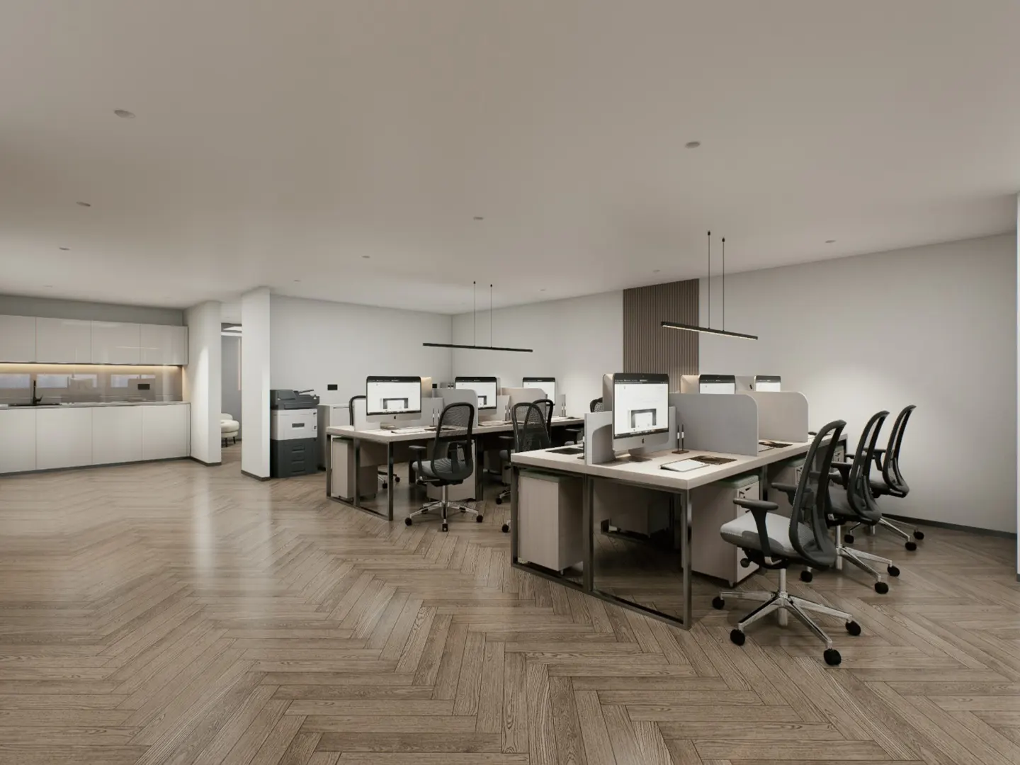 Bright office space with herringbone wood floors, desks with computers, and black chairs. A kitchen area is visible in the background.