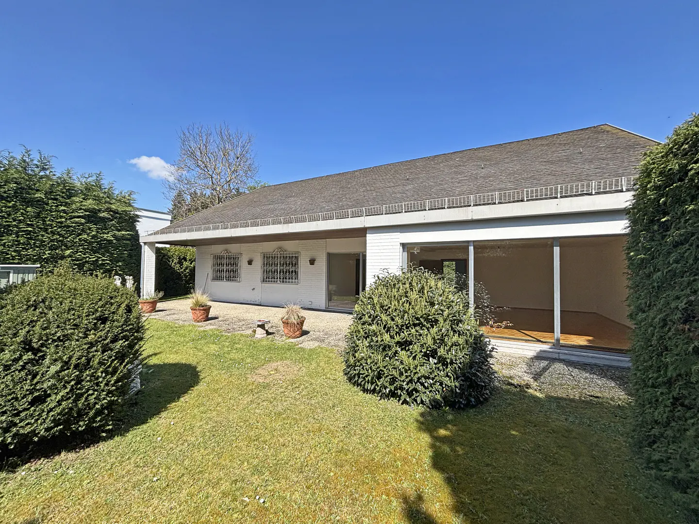 Exterior view of a one-story white house with a gray roof, green lawn, and trimmed hedges under a blue sky.