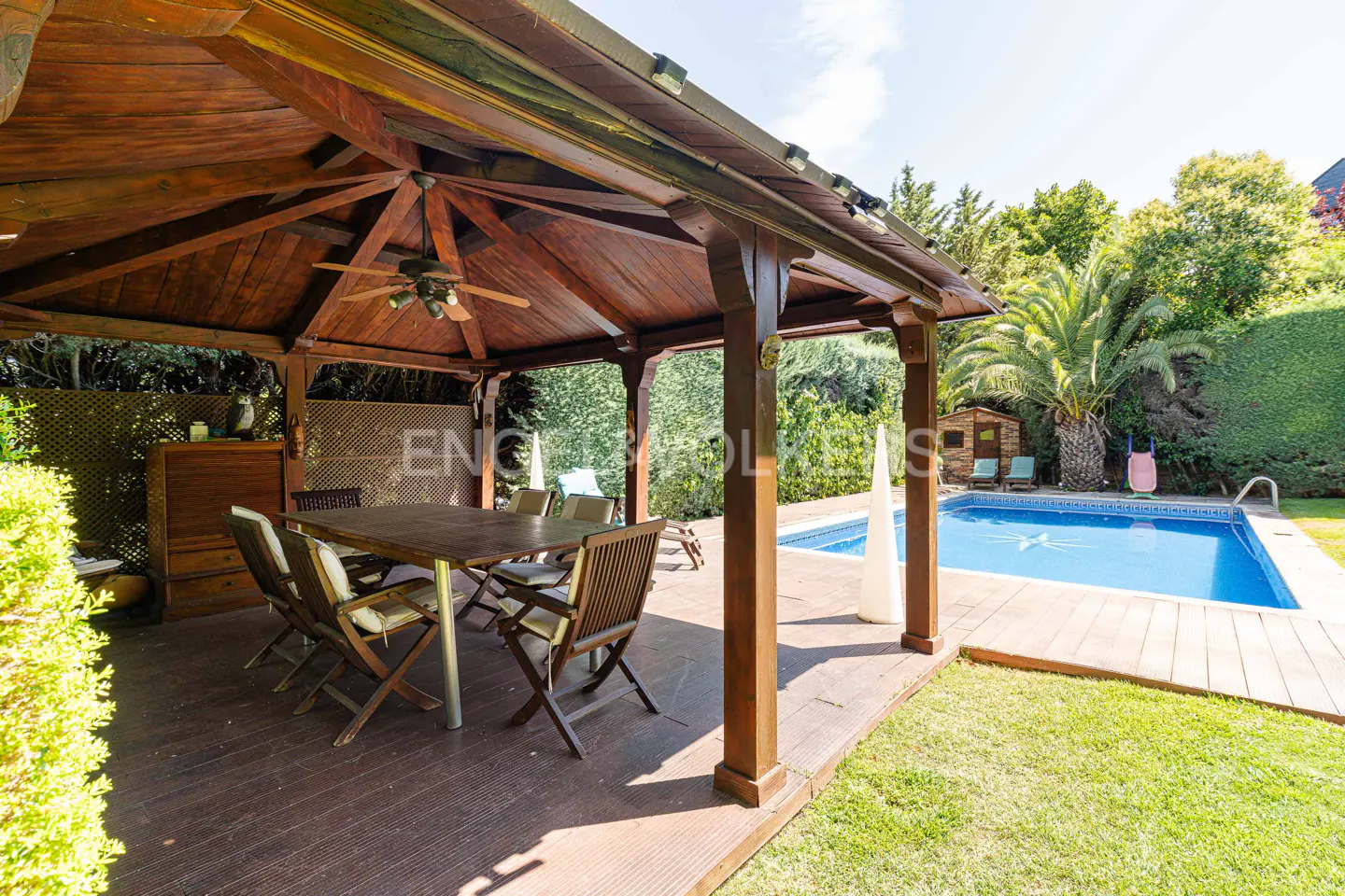 A backyard with a wooden gazebo, table, and chairs, next to a blue swimming pool and green lawn.