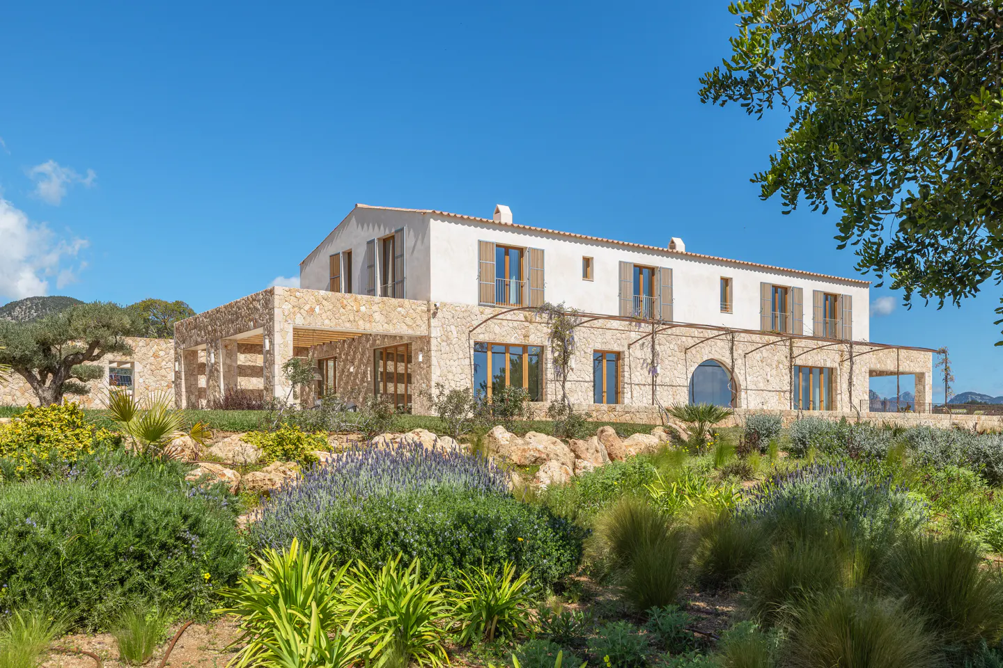 Two-story house with white walls, stone accents, and brown shutters, surrounded by lush green landscaping under a clear blue sky.
