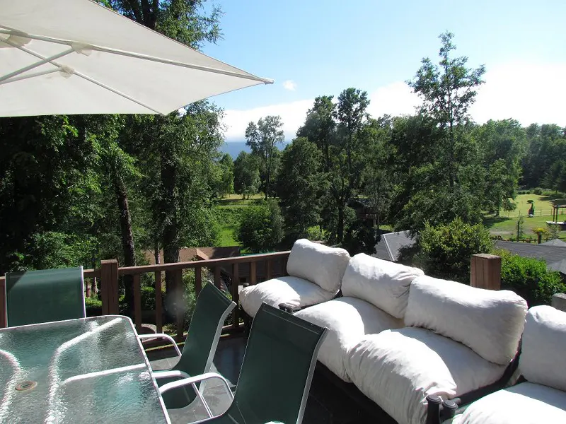 Outdoor patio with a glass table, green chairs, and a white cushioned sofa. A white umbrella provides shade, overlooking a lush green landscape.