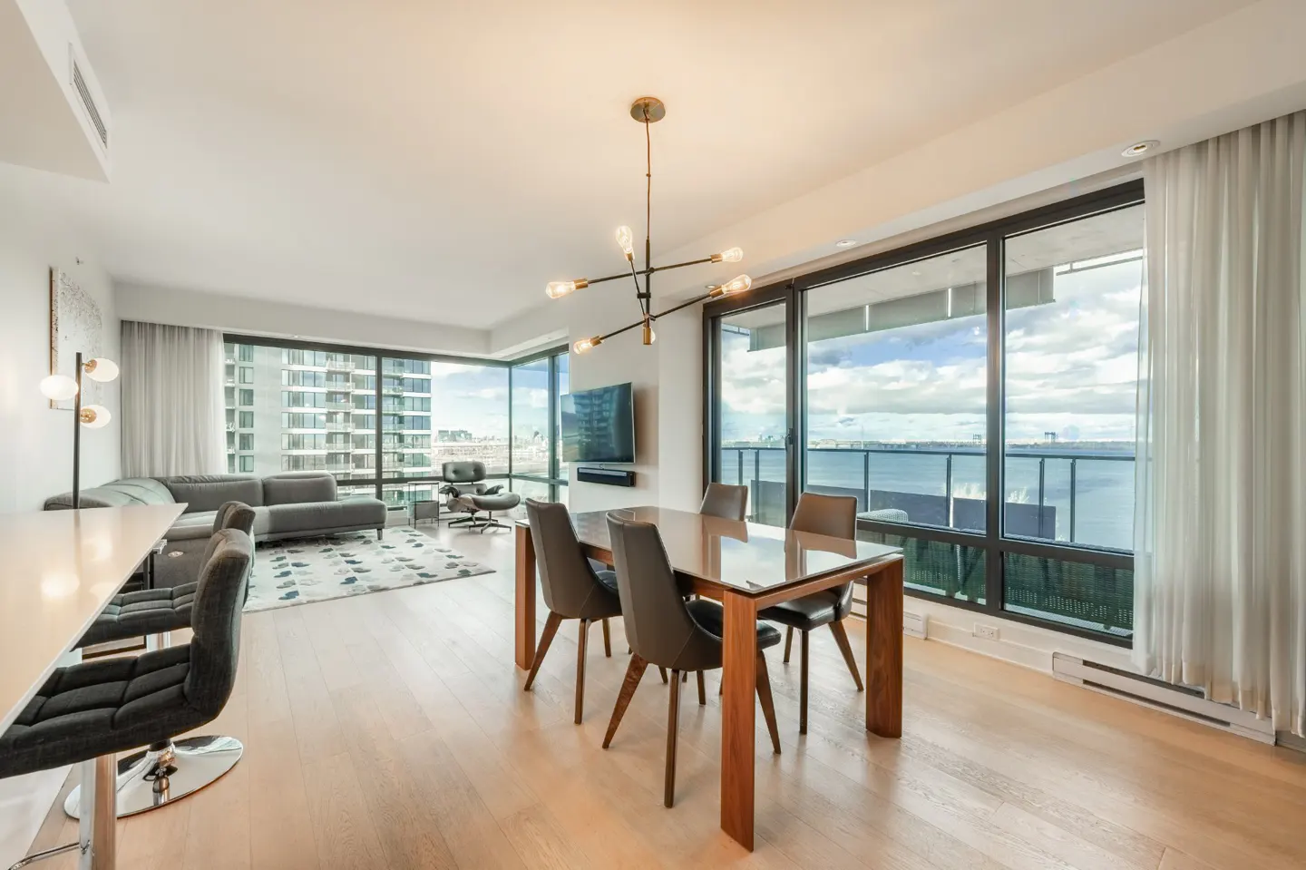 Bright, modern condo interior with wood floors, dining table, and floor-to-ceiling windows overlooking a city and ocean view.