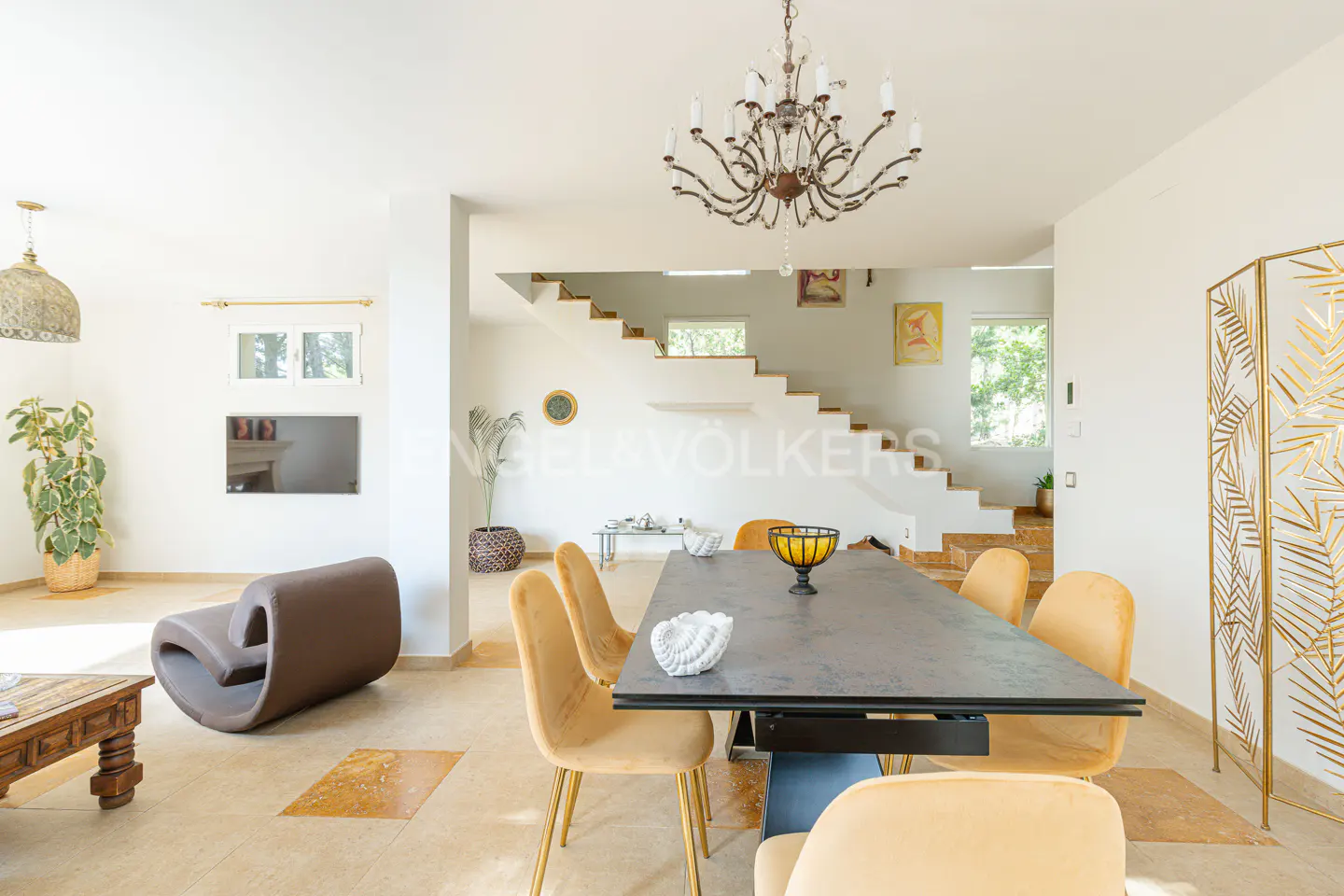 Bright, open-plan living and dining area with a large table, yellow chairs, and a decorative chandelier. Stairs lead to an upper level.