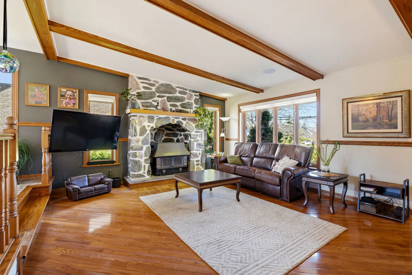 Living room with hardwood floors, stone fireplace, brown leather sofa, and a white patterned rug.