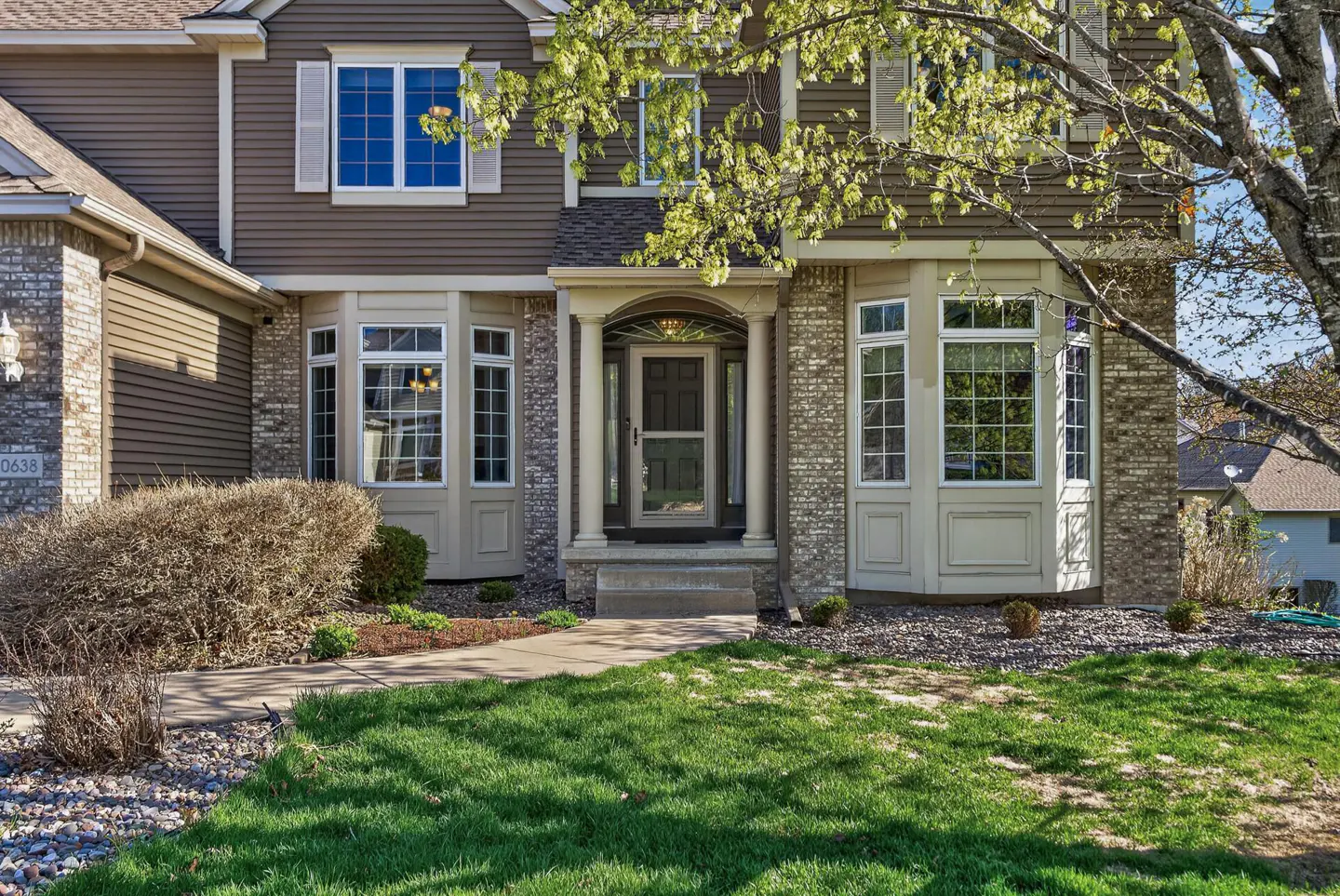 Two-story house with brown siding and brick accents. Bay windows flank the front door, which is framed by columns. Green lawn in the foreground.
