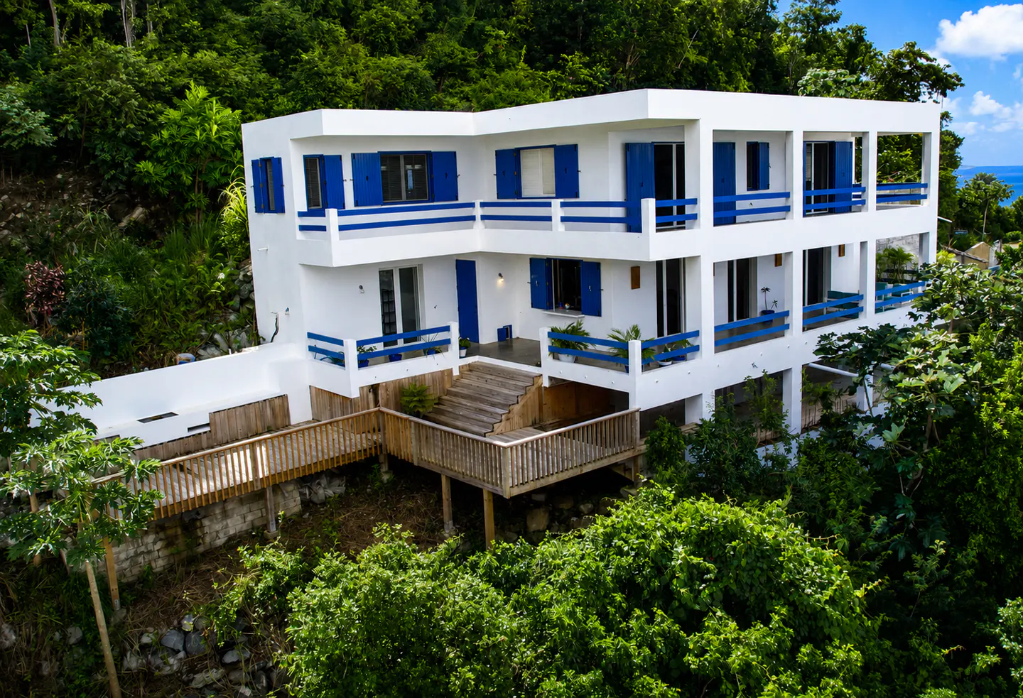A three-story white house with blue shutters and railings, nestled in lush green trees. A wooden walkway leads to the house.