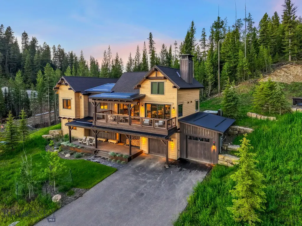 A tan two-story house with a black roof, a deck, and a garage, surrounded by green trees.