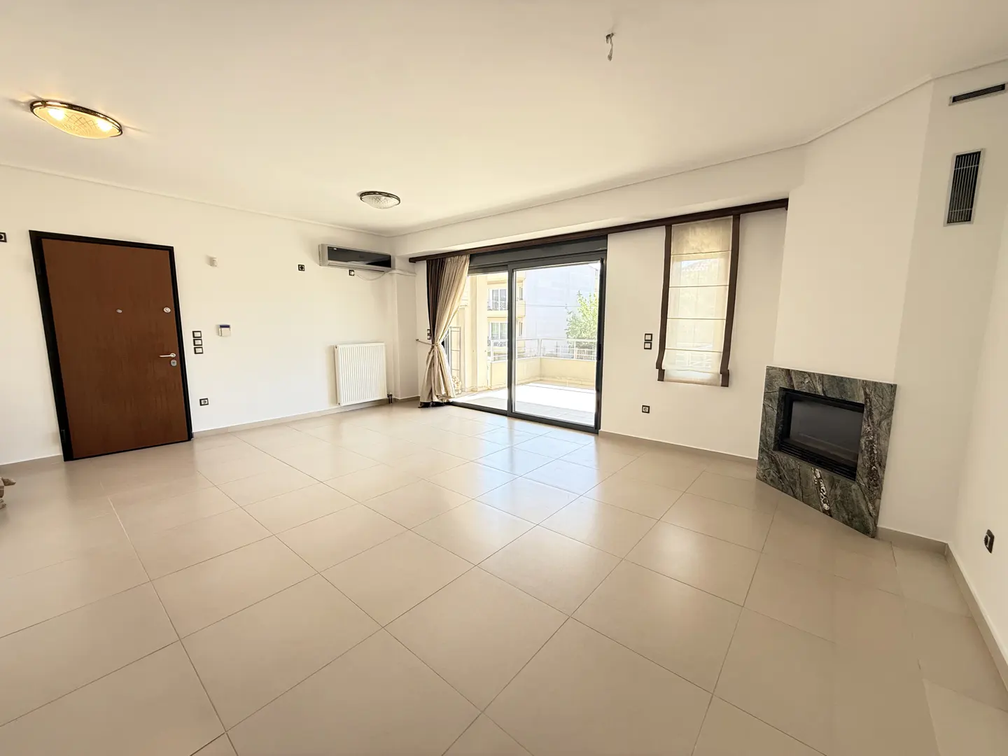 Bright, empty living room with beige tile floor, white walls, brown door, fireplace, and sliding glass doors to a balcony.