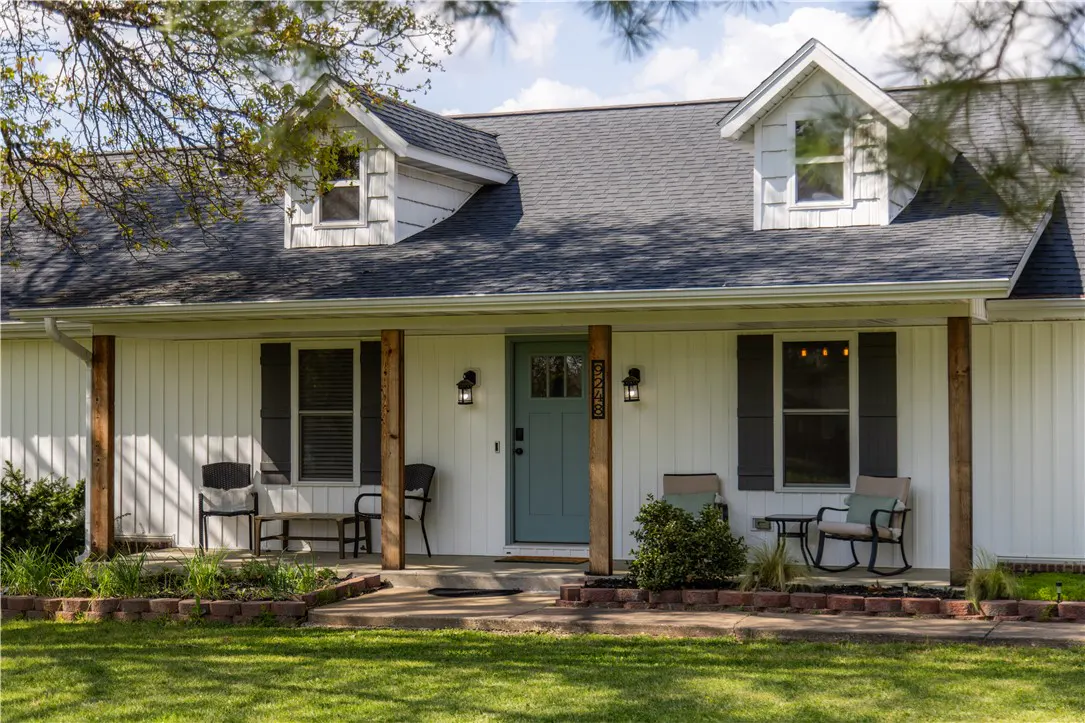 Exterior of a white house with a gray roof, dormers, and a blue front door. The porch has wooden pillars and seating.