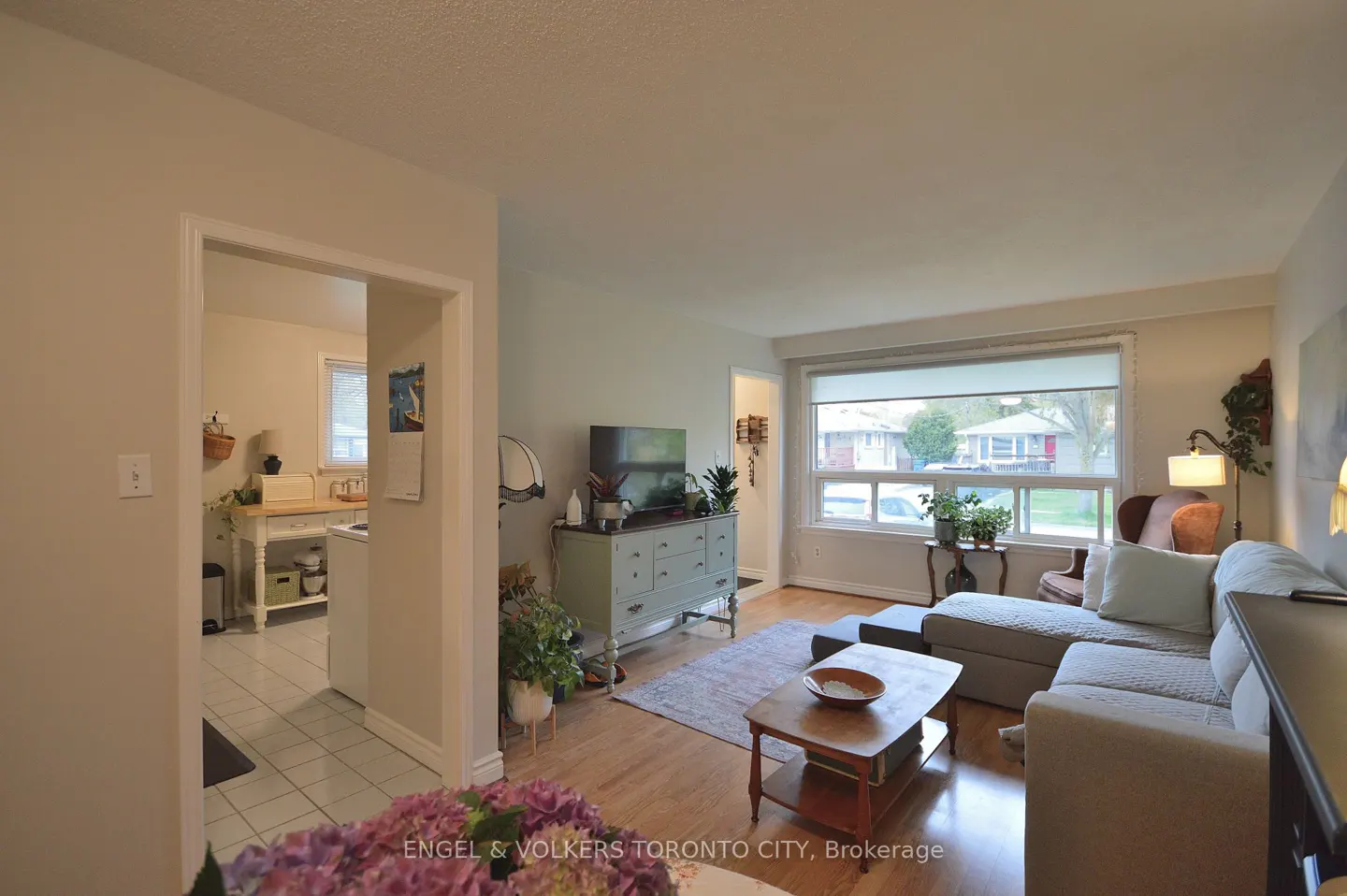 A bright living room with a gray sofa, wooden coffee table, and a large window. A kitchen is visible through an open doorway.