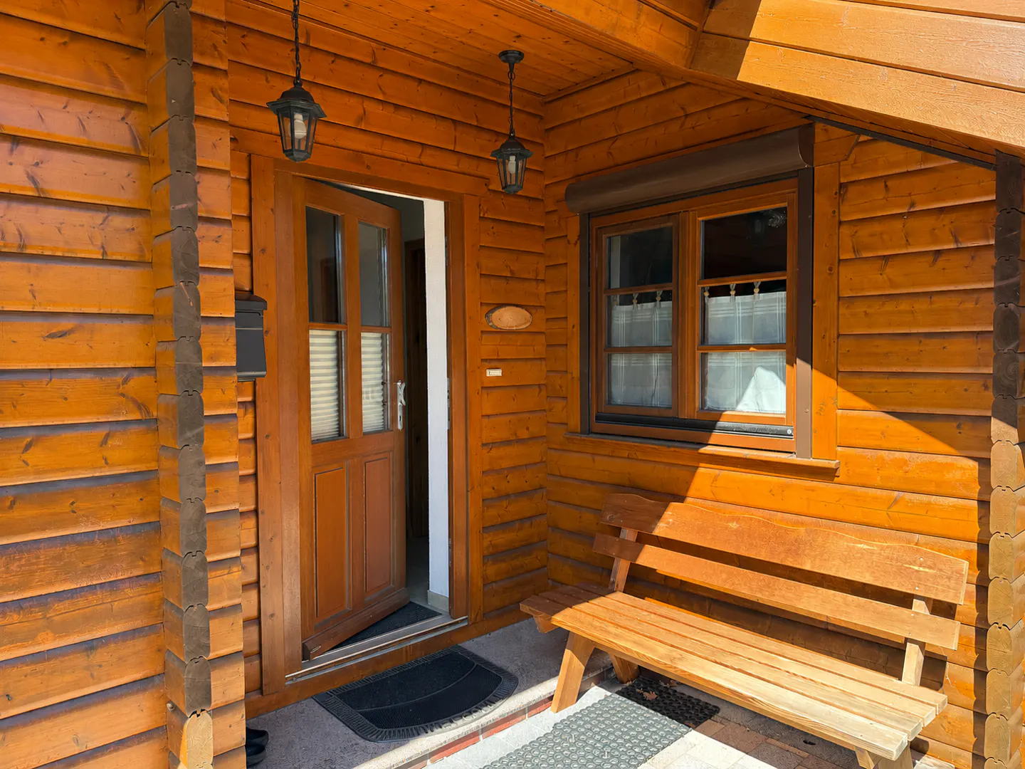 Exterior view of a wooden cabin with an open door, a window, a bench, and hanging lanterns.