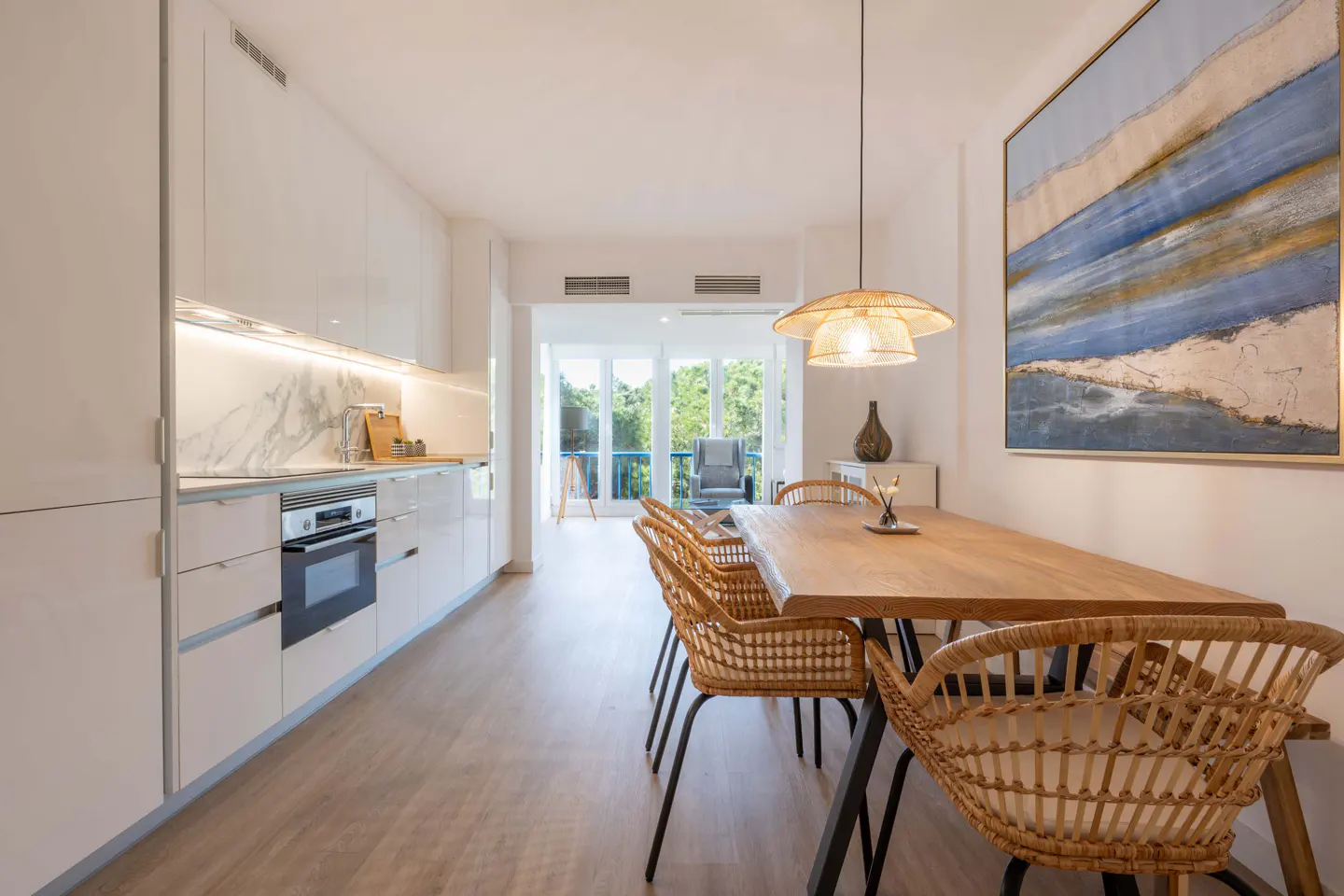 Bright, modern kitchen and dining area with white cabinets, wood table, wicker chairs, and blue abstract art. Balcony in background.