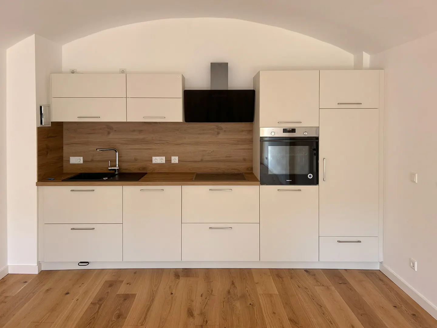 A modern kitchen with white cabinets, wood countertops, and a black range hood. The floor is wood.