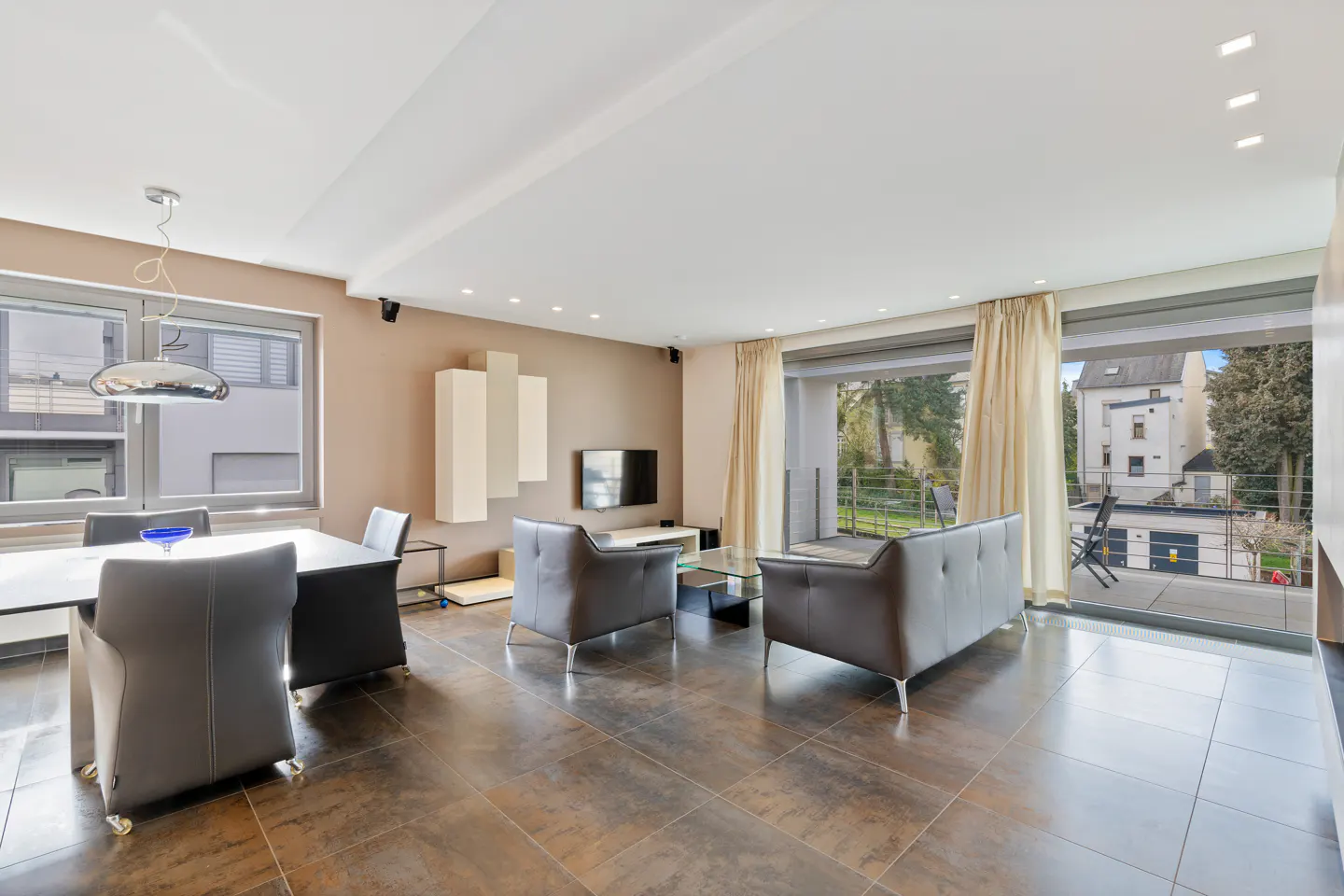A modern living room with a dining table, gray chairs, and a gray sofa. Large windows offer a view of the outdoors.