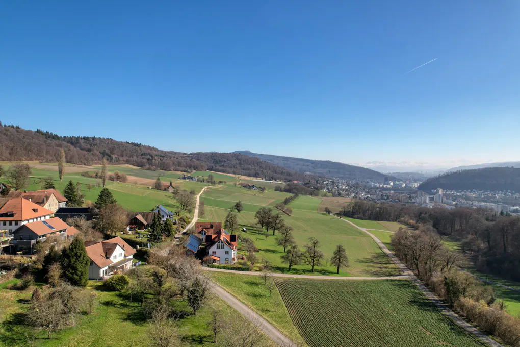 Aerial view of houses with red roofs in a green, hilly landscape under a clear blue sky.