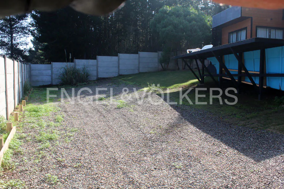 Gravel driveway leading to a modern house with a blue carport, surrounded by a gray concrete fence and green trees.