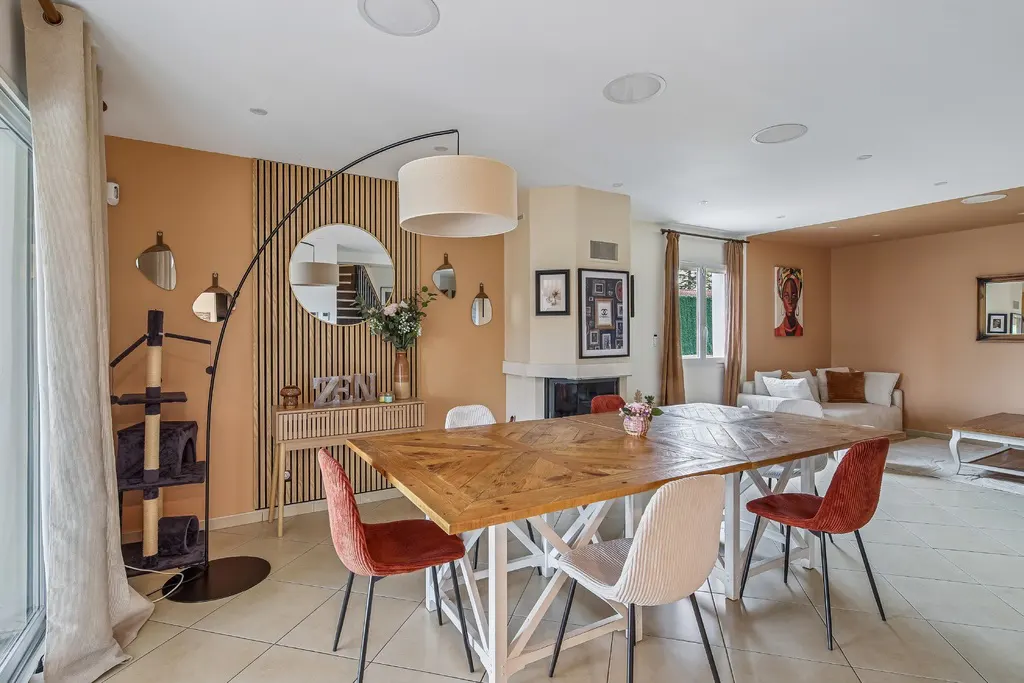 A bright, open-concept dining and living room with a large wooden table, fireplace, and modern decor. The walls are painted in warm, earthy tones.