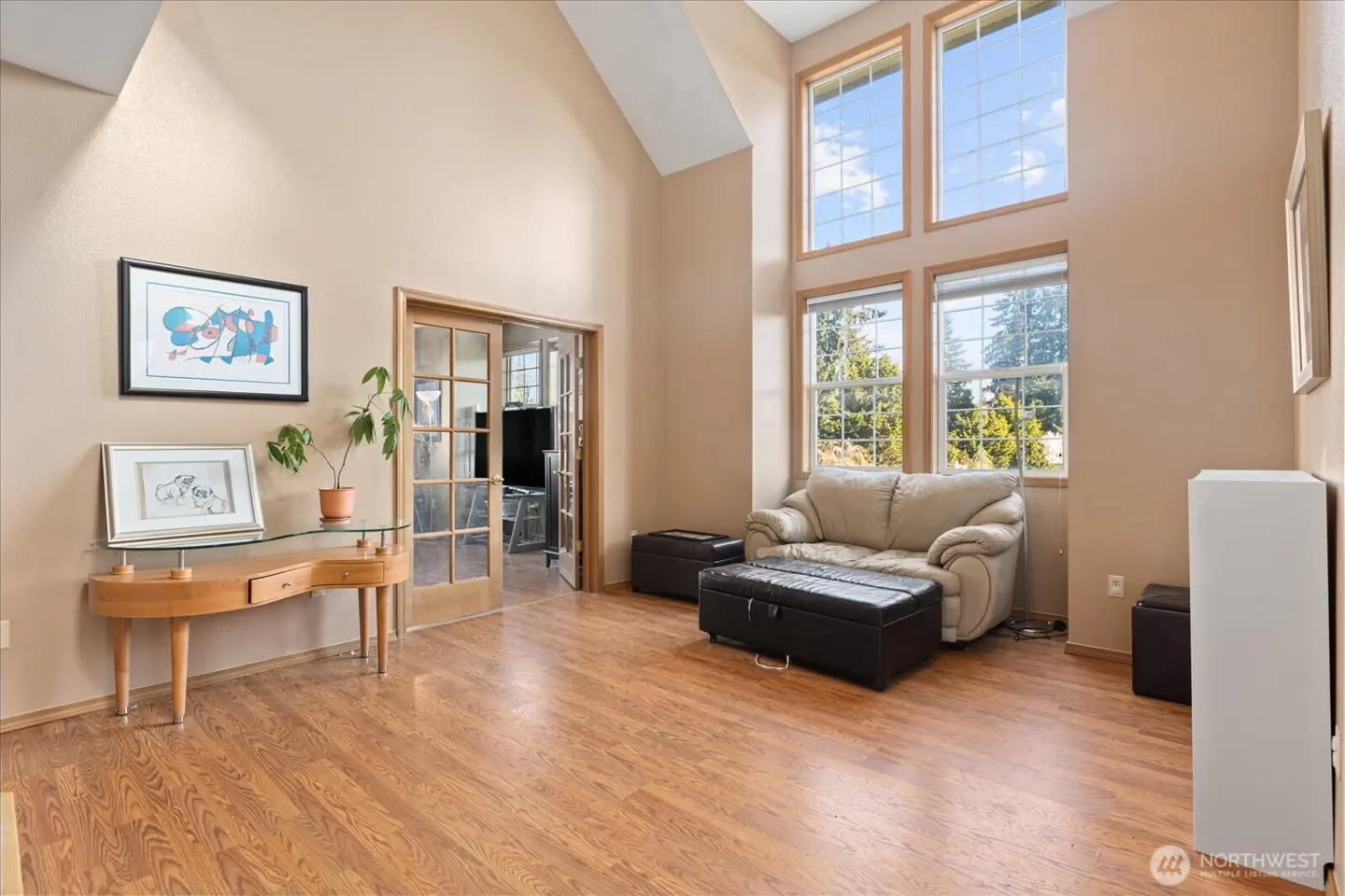 Living room with wood floors, beige walls, and tall windows. A beige sofa and black ottoman sit near the windows. A desk and plant are on the left.