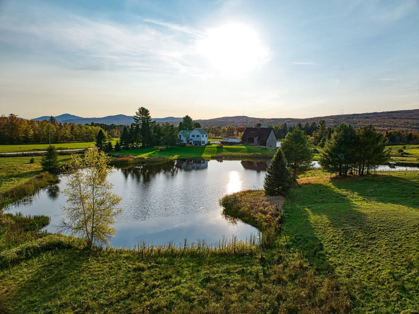 Scenic aerial view of a pond reflecting a house and barn under a sunny sky, surrounded by green fields and trees.