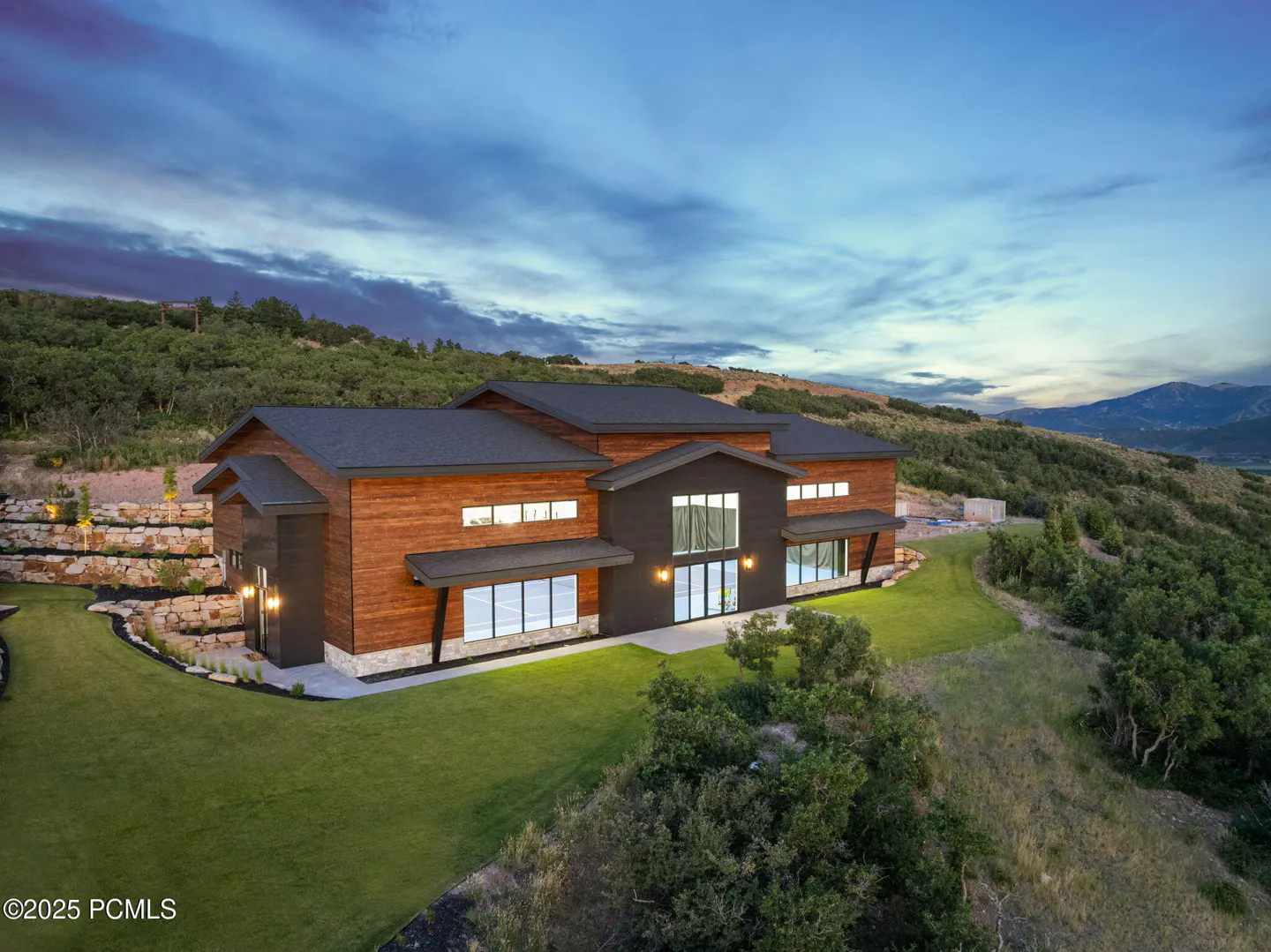 Modern mountain home with wood siding and large windows, set on a grassy hillside at dusk under a blue sky.
