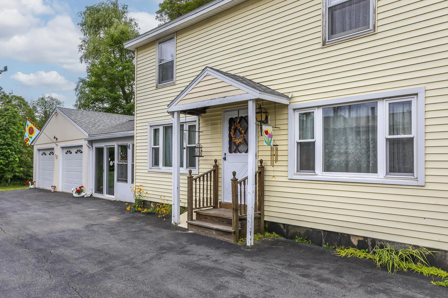 Two-story yellow house with white trim, a covered porch, and a detached garage with a sunflower flag.