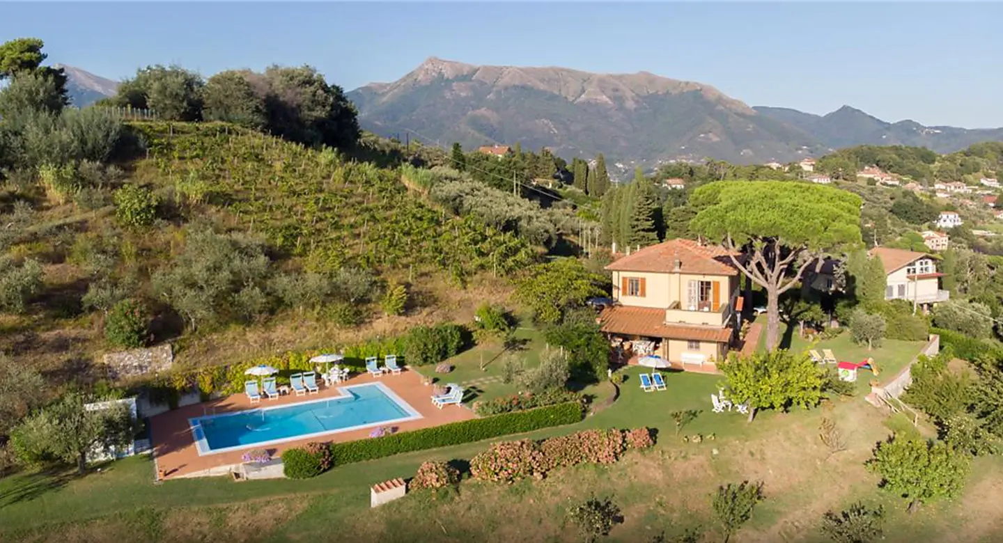 Aerial view of a tan villa with a red tile roof, a pool, and a mountain backdrop.
