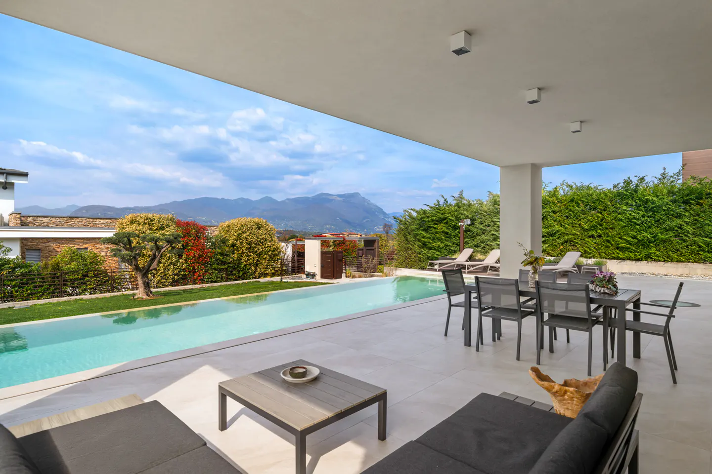 Outdoor patio with a pool, table, and chairs. Mountains and blue sky are in the background.
