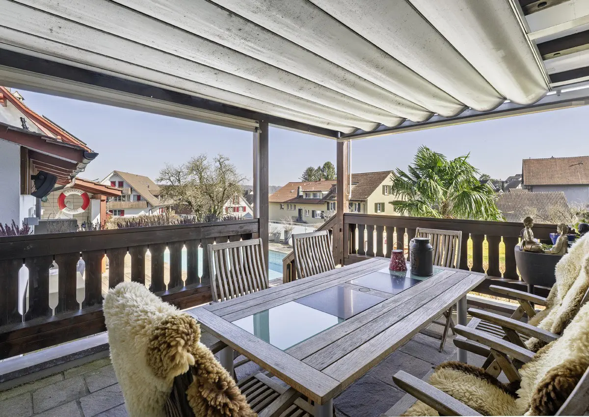 Covered patio with a wood table, chairs with sheepskin throws, and a view of houses and a pool.