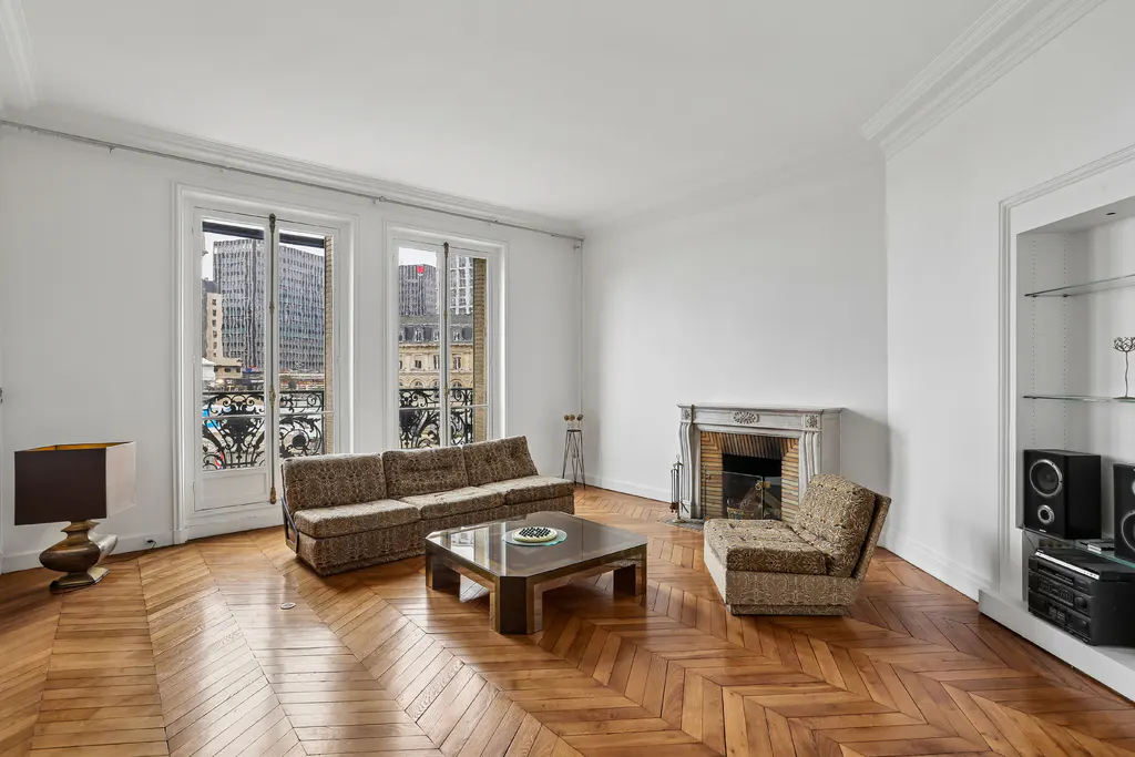Bright living room with herringbone wood floors, patterned sofa and chair, fireplace, and city view through balcony doors.