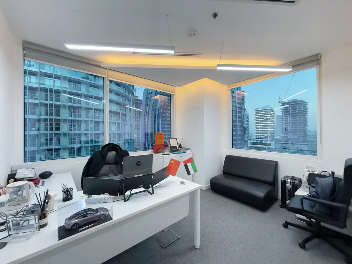 Bright office with a white desk, black sofa, and large windows showing city views. Flags of China and UAE are on the desk.
