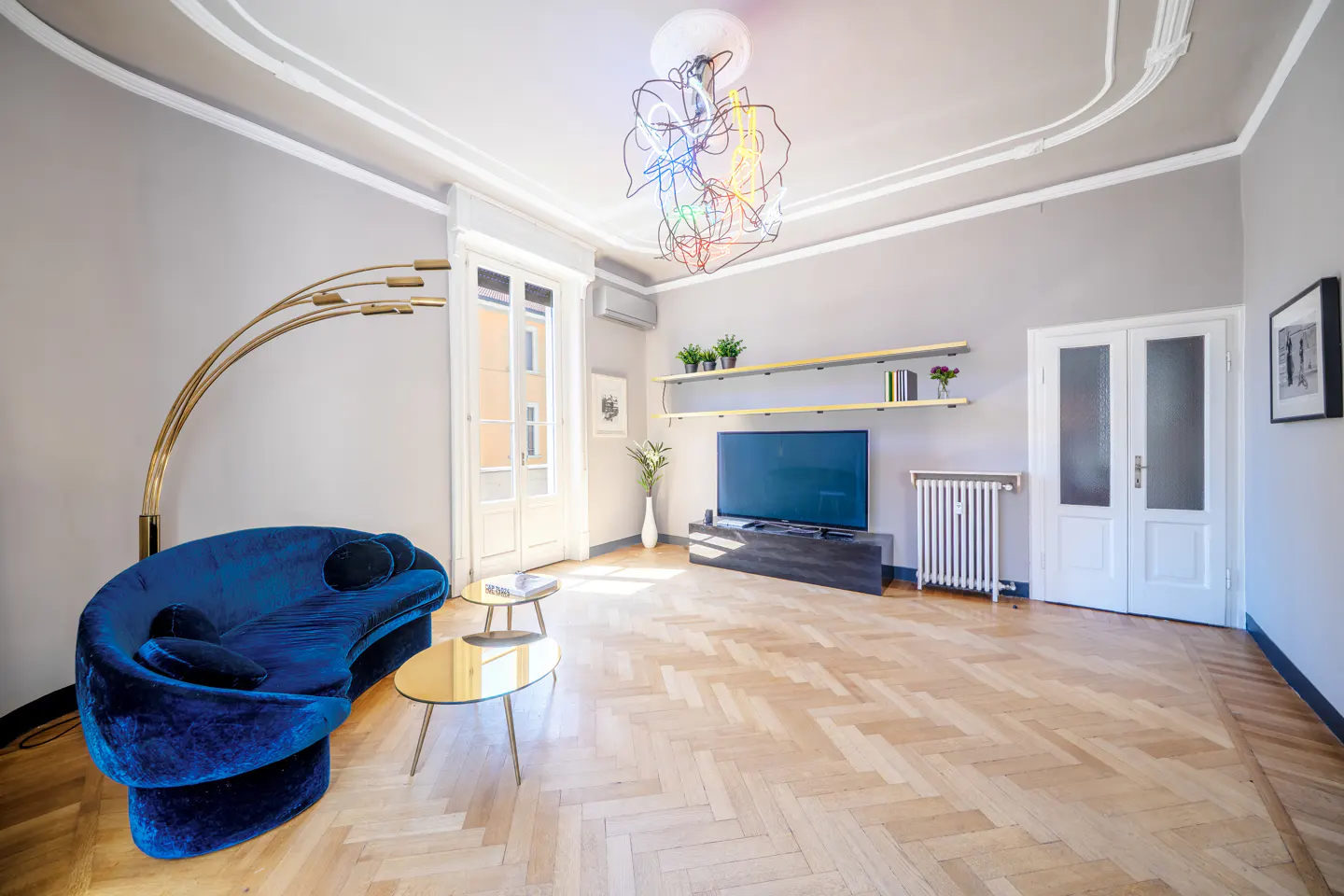 Bright living room with herringbone wood floors, blue velvet sofa, gold accents, and a modern chandelier.