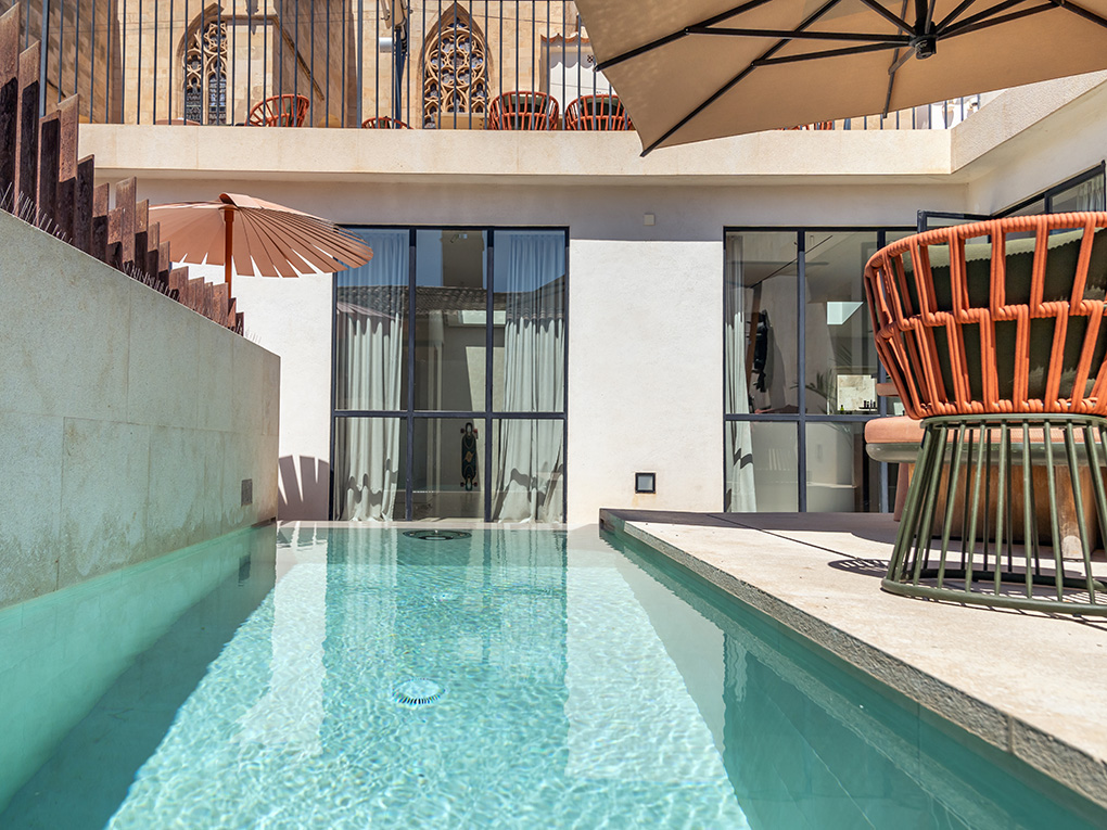 A modern patio with a turquoise plunge pool, orange woven chairs, and a beige umbrella against a historic stone building. [AI-generated text]