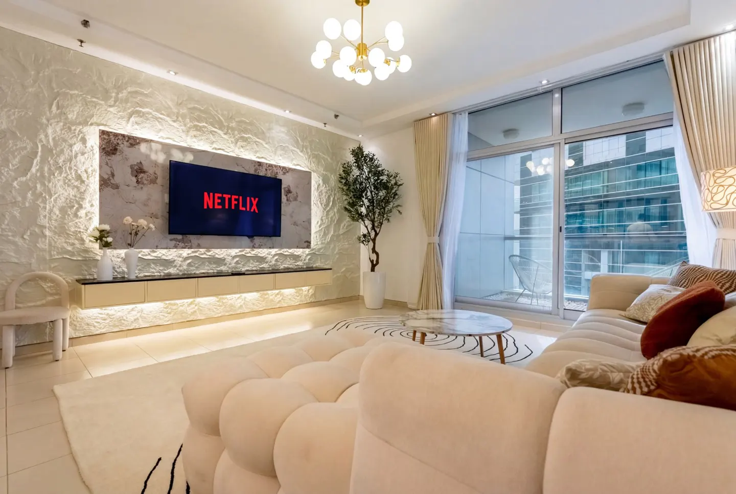 Living room with a large cream sofa, marble table, and a TV with Netflix on a textured wall. A modern chandelier hangs from the ceiling.