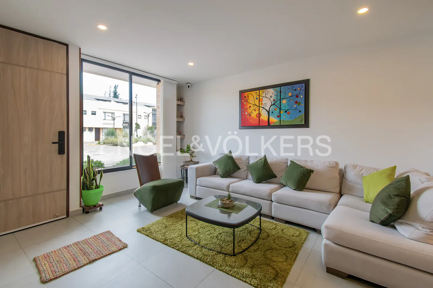 A modern living room with a beige sectional sofa, green pillows, and a colorful tree painting. A green rug and chair add pops of color.