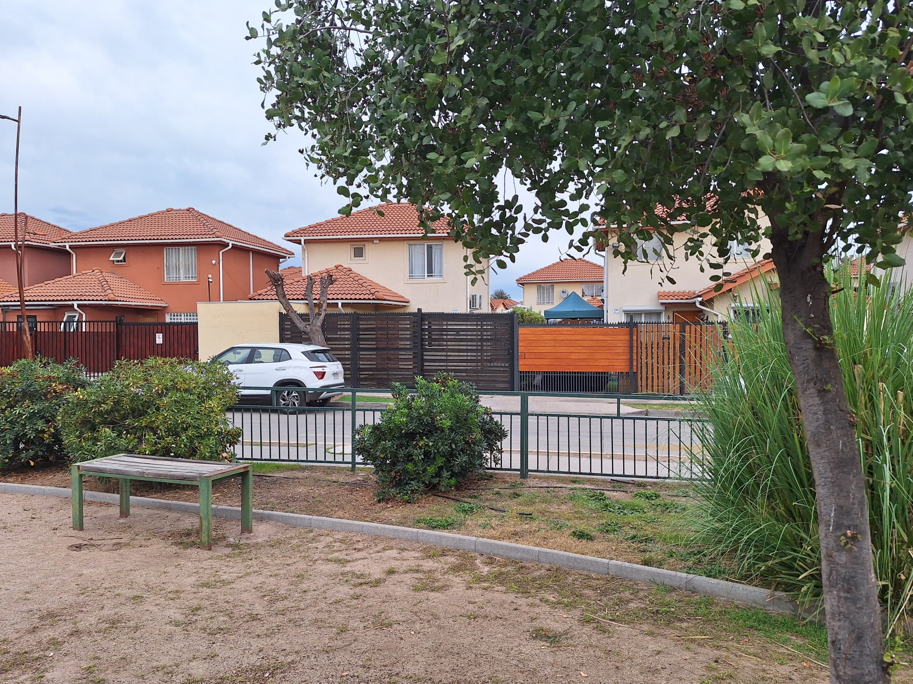 A white SUV parked behind a metal fence in front of suburban houses with orange tiled roofs and a small wooden bench.
