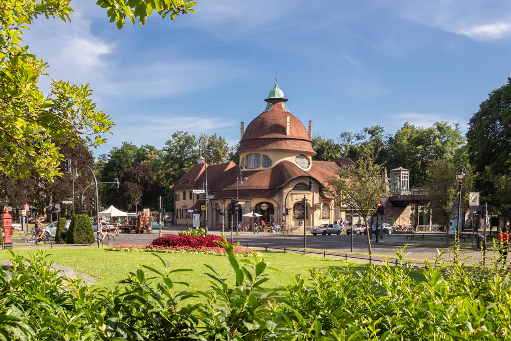 A park view of a building with a red dome roof, green lawn, trees, and people on bikes.