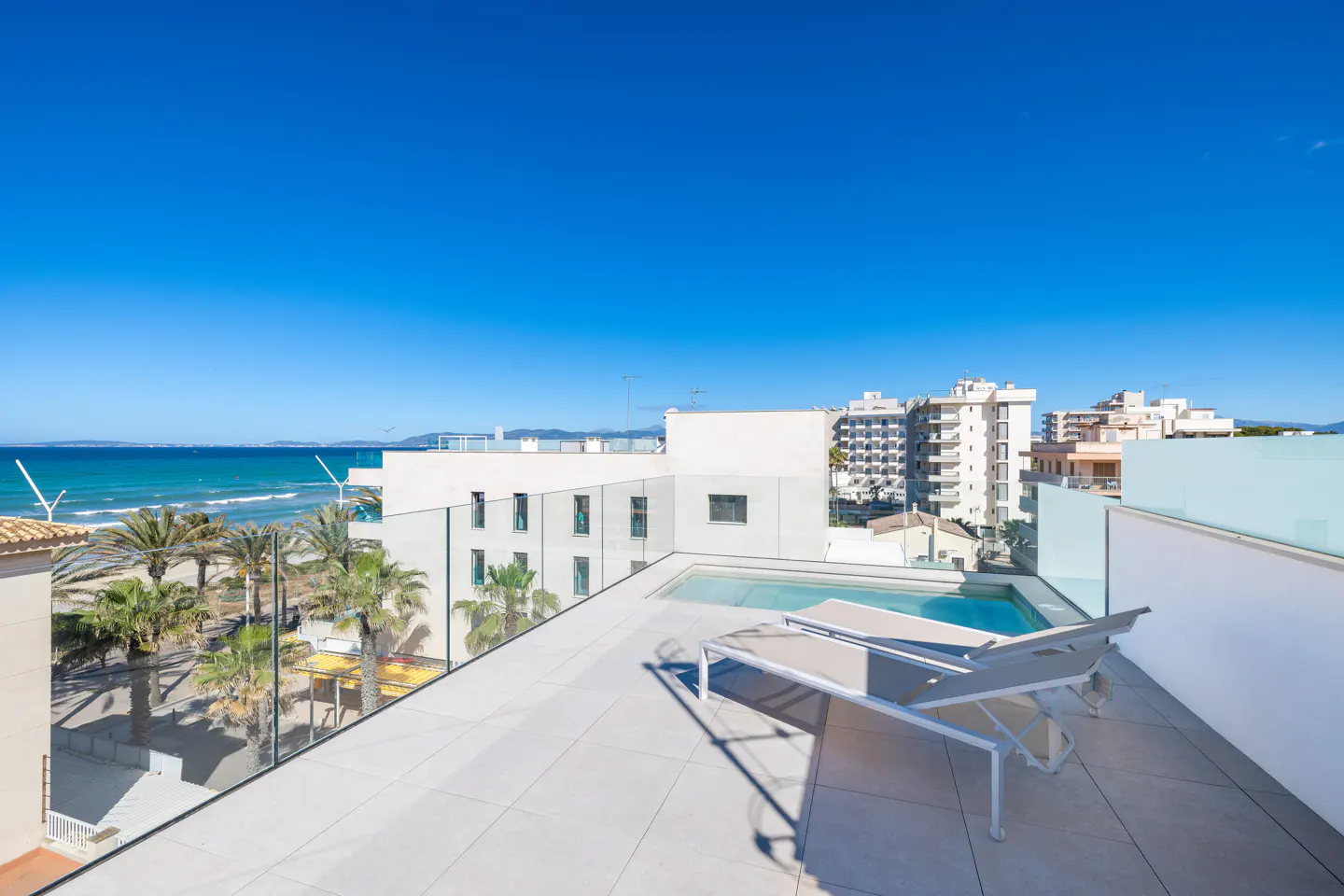 Rooftop view of a pool and lounge chair with a beach and buildings in the background on a sunny day.