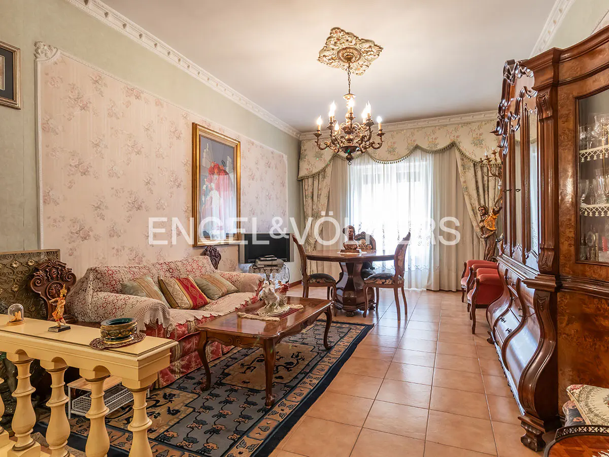 Ornate living room with floral wallpaper, chandelier, sofa, and dark wood furniture. A round table sits near the window.