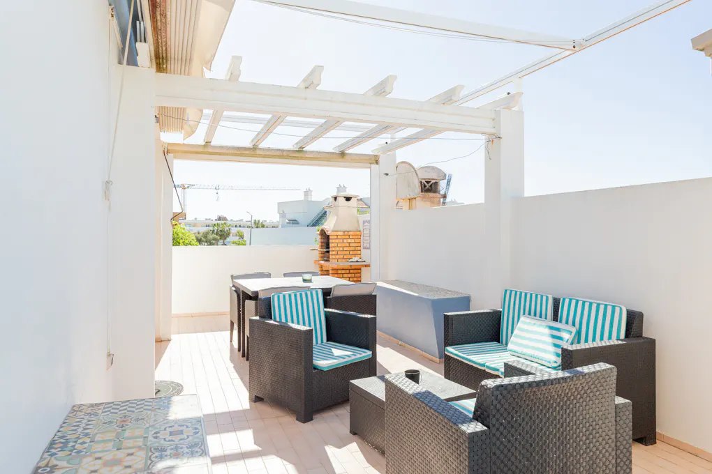 Outdoor patio with black wicker furniture, blue striped cushions, and a white pergola. A brick grill is visible in the background.