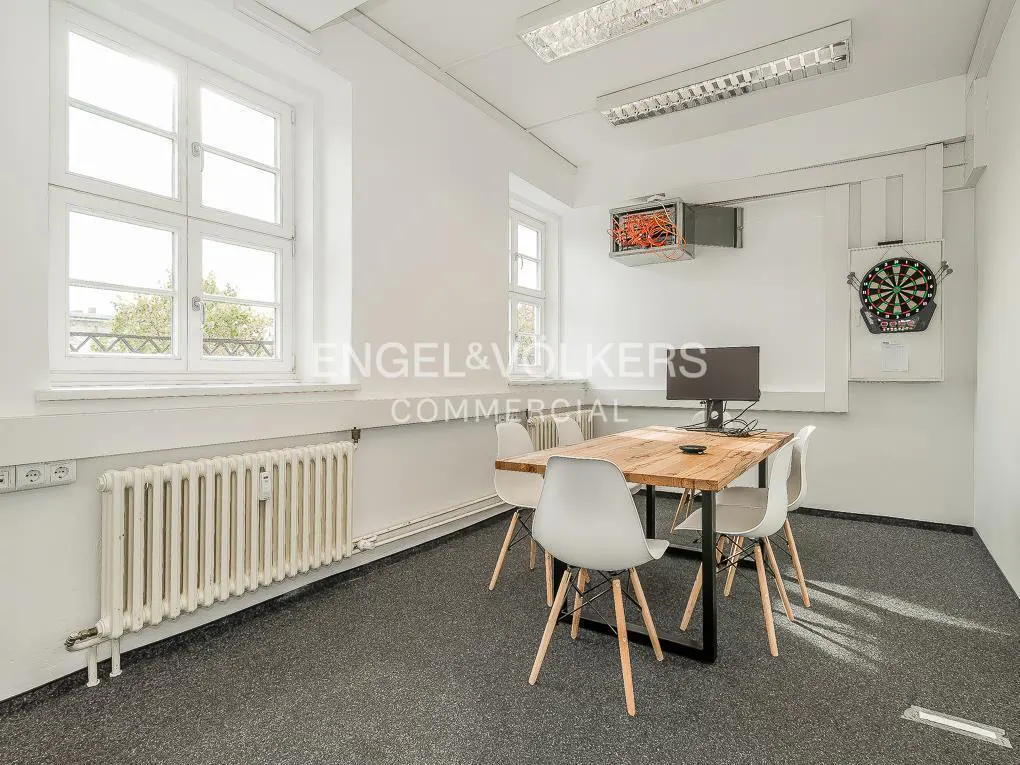 Bright office space with a wooden table, four white chairs, a computer, and a dartboard on the wall.