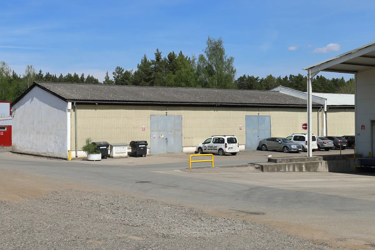 Exterior of a long, one-story warehouse with gray doors and a parking lot with several cars.