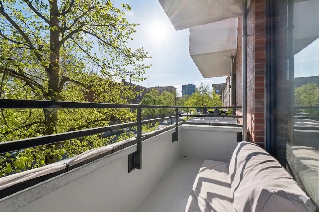 Apartment balcony with black metal railing overlooks a tree-lined street. A covered sofa sits on the white floor. Brick building on the right.