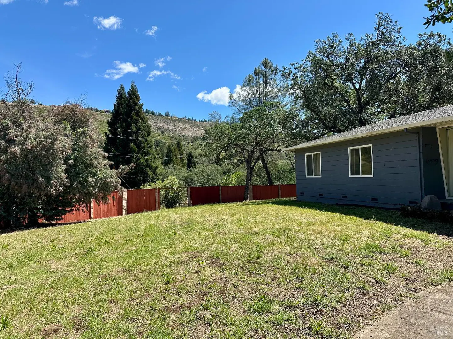 Exterior view of a gray house with a red fence, green lawn, trees, and a mountain in the background under a blue sky.