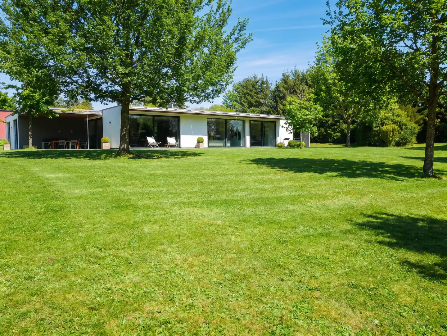 Modern white house with large windows, surrounded by green lawn and trees under a blue sky. Two white chairs sit outside.