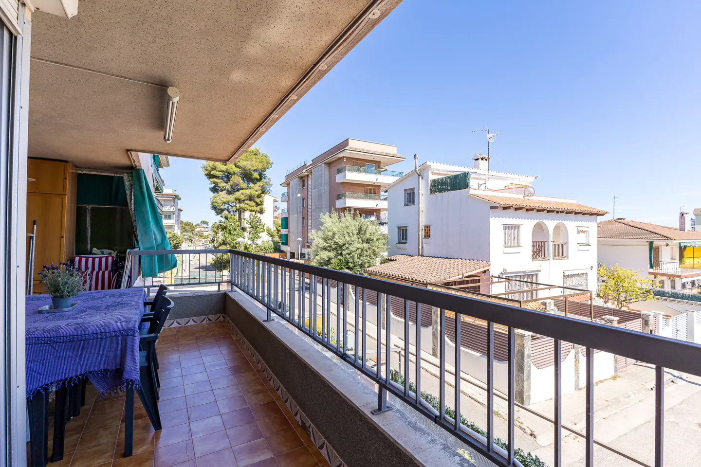A balcony with a table and chairs overlooks a street with houses under a blue sky.