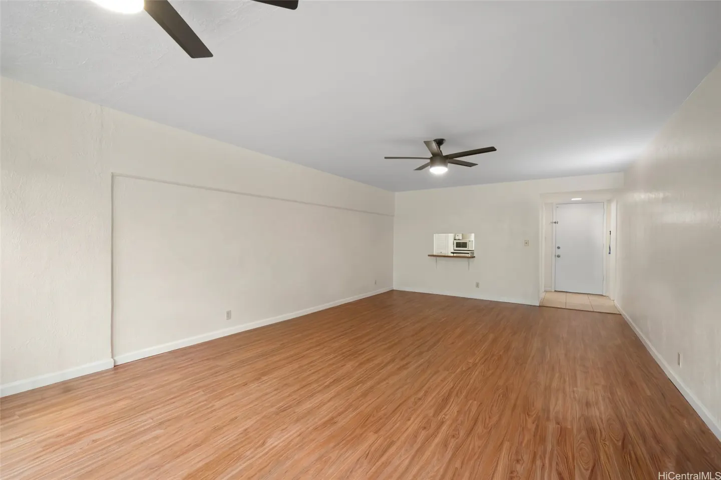 A bright, empty living room with wood floors, white walls, and two ceiling fans. A pass-through to the kitchen is visible.