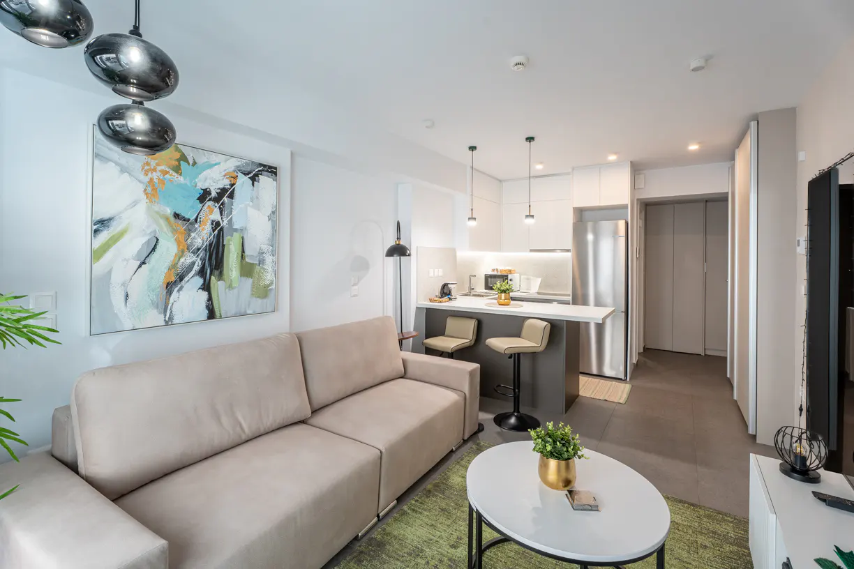 Modern studio apartment with a beige sofa, white oval coffee table, and a kitchen island with two bar stools.