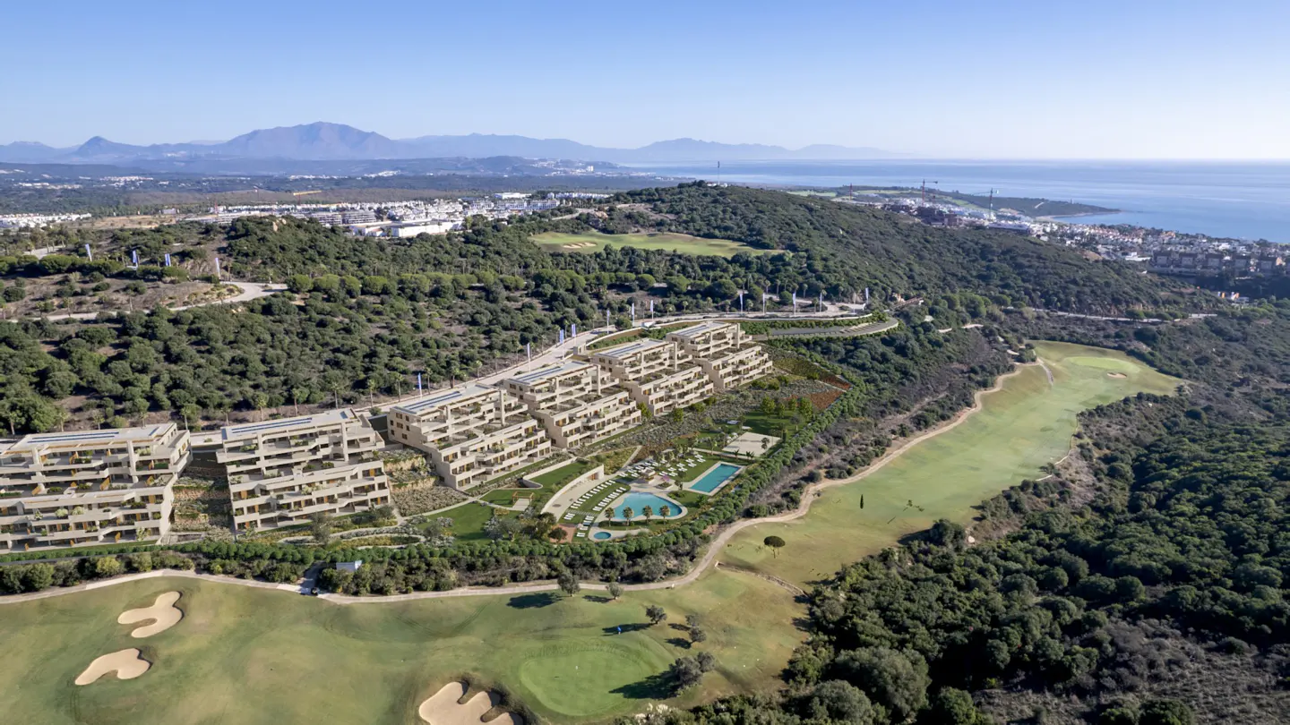 Aerial view of modern apartments with pools on a green hillside near a golf course and the ocean.